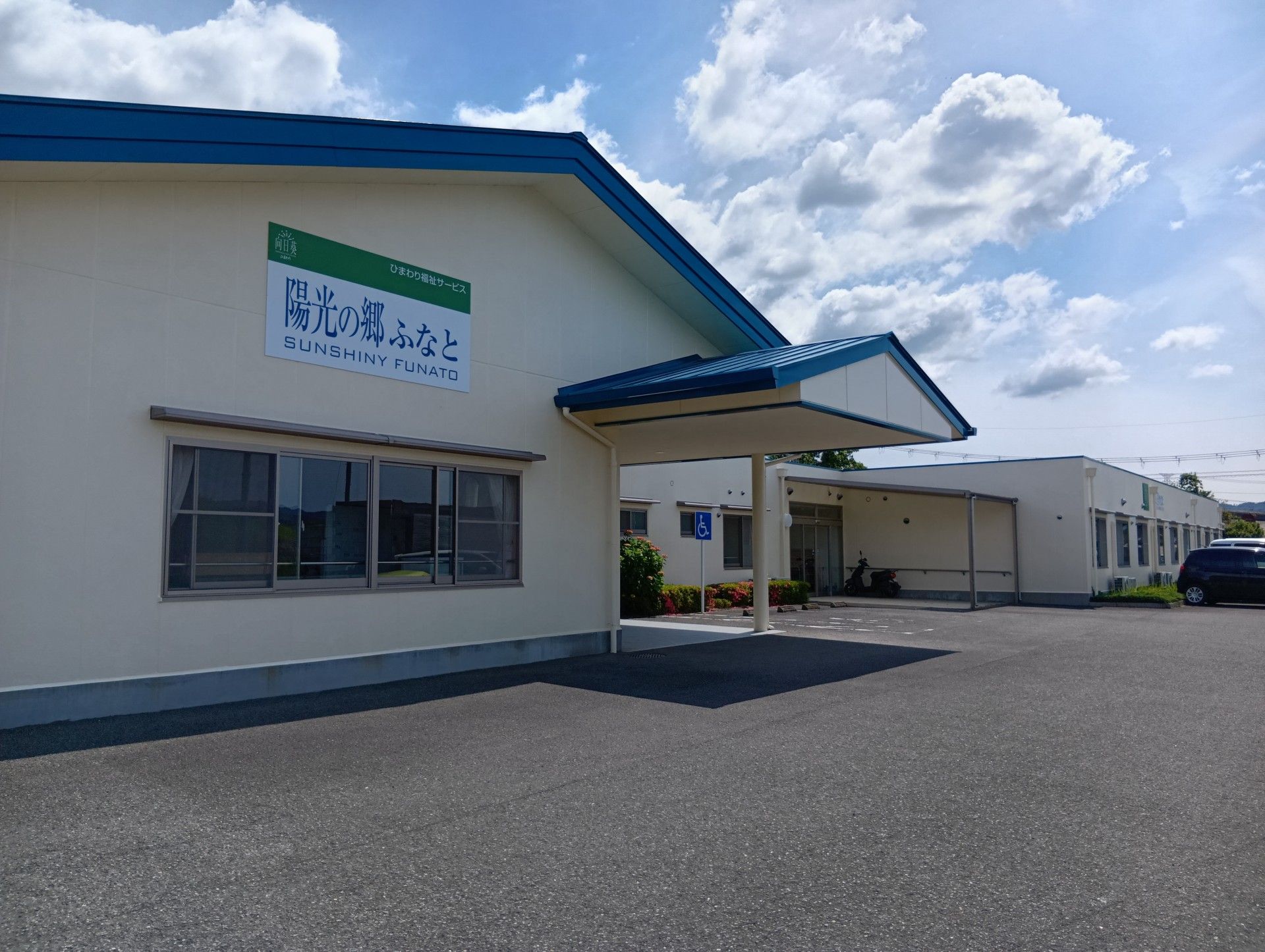 緑の多い屋外で笑顔で過ごしている車いすの高齢女性と付き添いのピンクのエプロンを付けた若い女性スタッフ。
