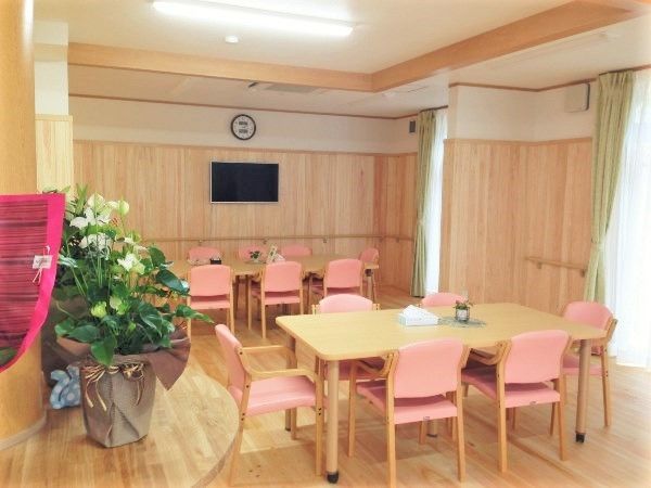 大きな壁掛けテレビが設置されたデイサービス。ダイニングテーブルが複数並んでいる。