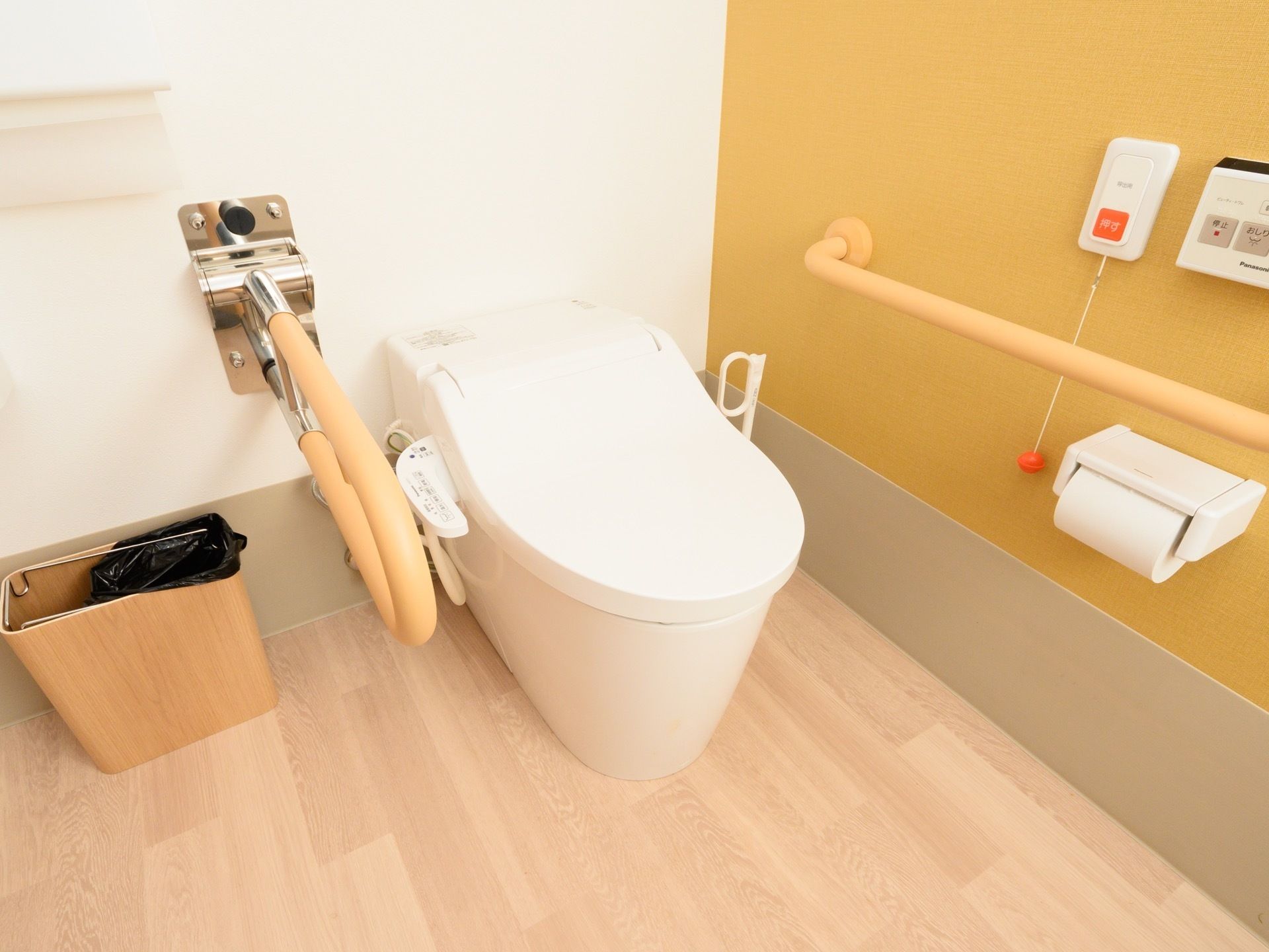 温水洗浄機能付きのトイレである。長い紐が付いた緊急時呼び出しボタンが備わっている。便器の両側に手すりが設置されている。