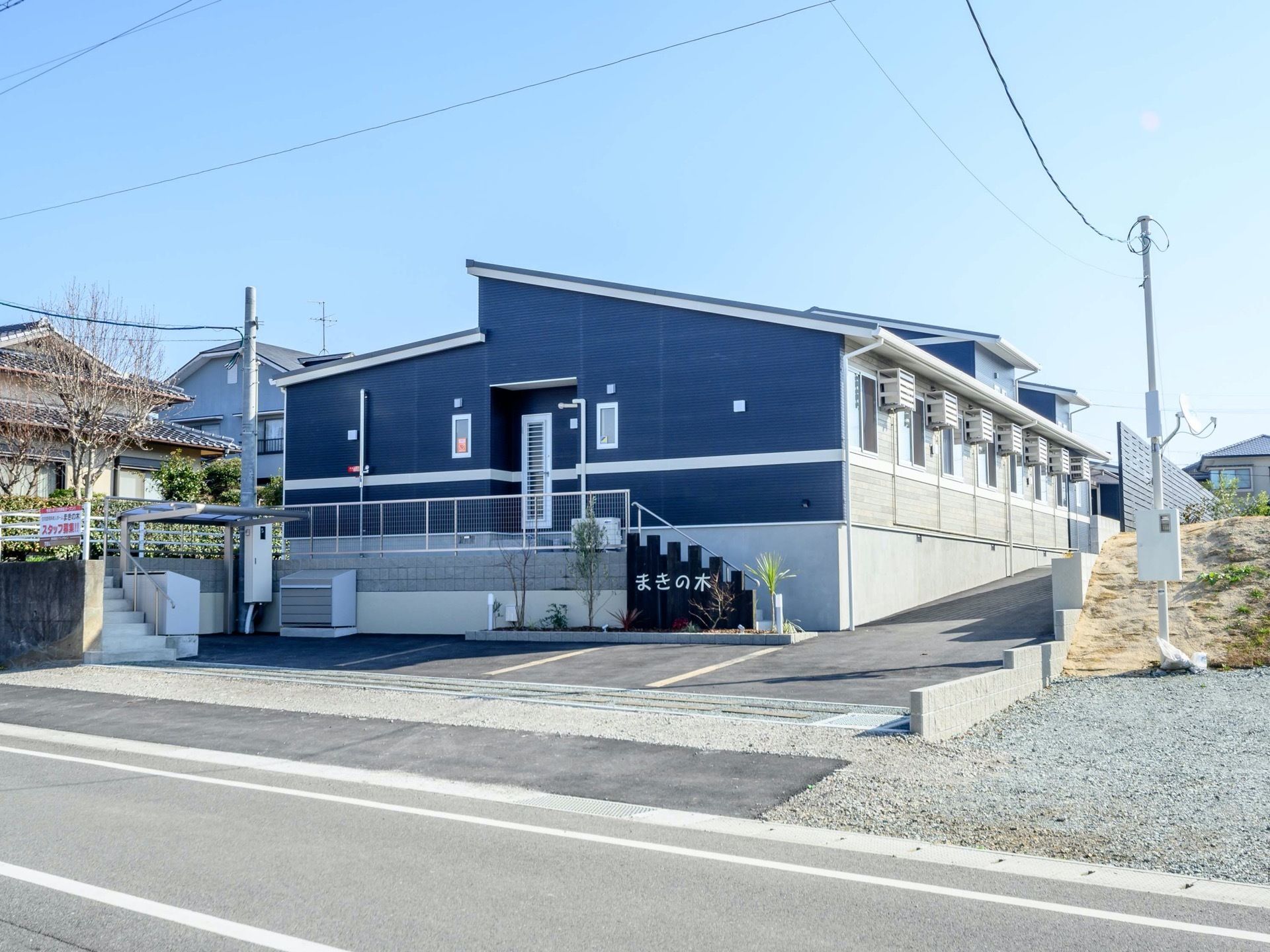 濃紺の壁に白い線が描かれた爽やかな印象の外観である。道路から正面には勝手口があり、正面玄関は奥まった場所にある。