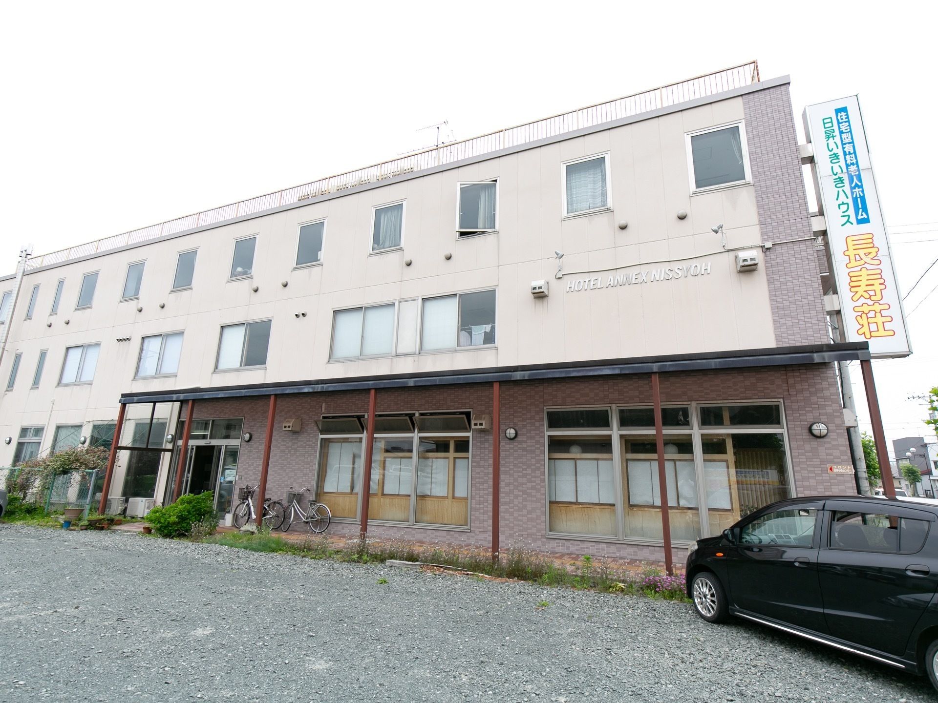 ３階建ての屋上のある建物。施設名が書かれた大きな看板が設置されている。正面部分は砂利が敷かれた駐車場がある。
