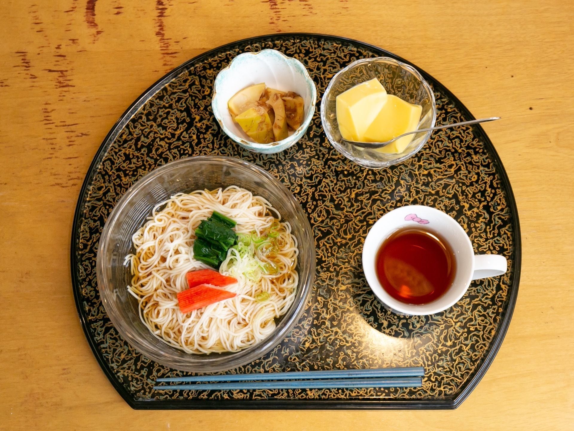 黒塗りに金の模様が描かれているお盆に食事が乗っている。素麺、和え物が入った小鉢、デザート、お茶が置かれている。