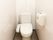 白い壁と茶色の床の個室のトイレがある。便座には蓋があり、温水洗浄便座が設置されている。壁にはペーターホルダーが付いている。