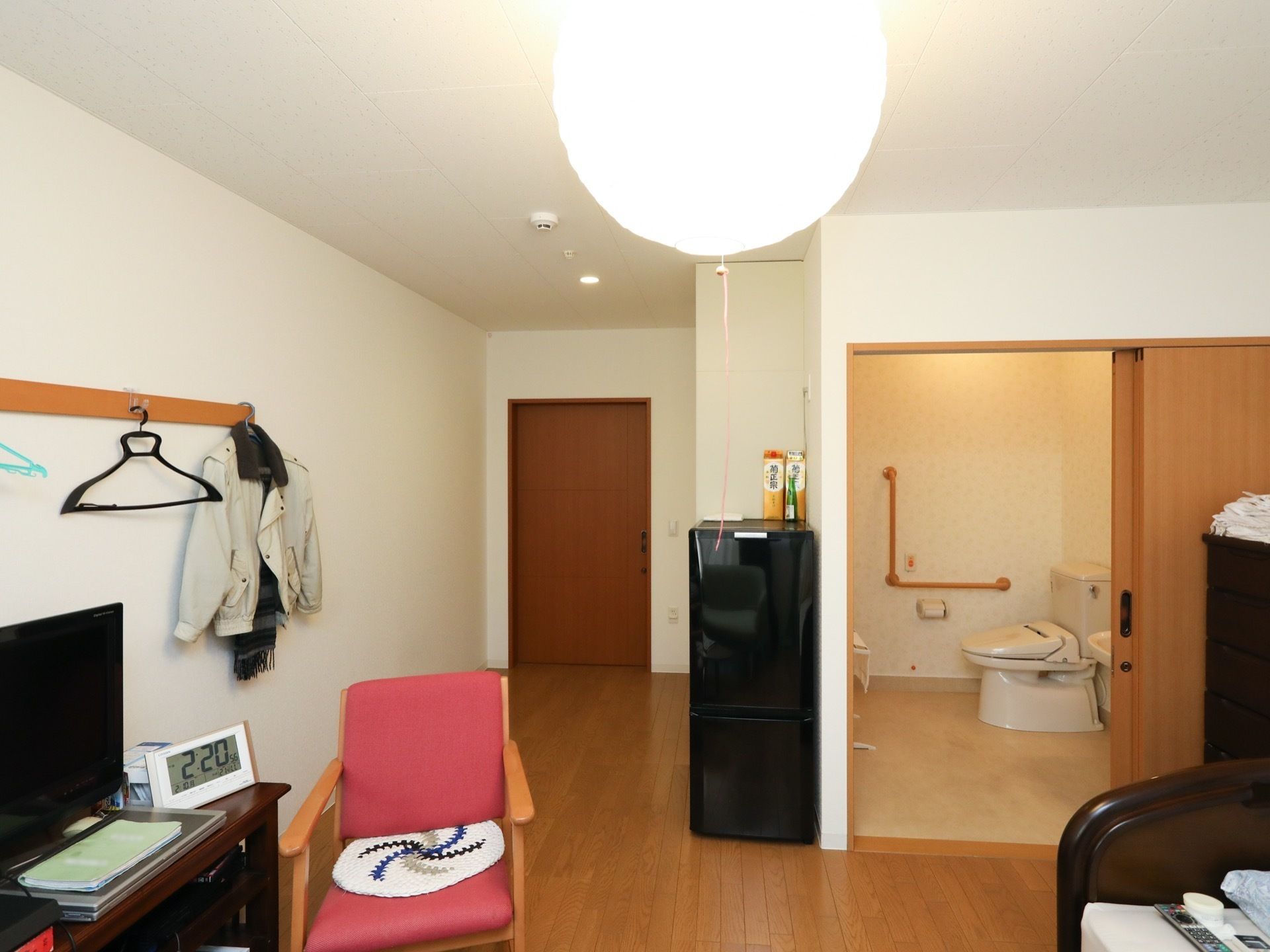居室内には冷蔵庫や箪笥、テレビ台などいくつかの家具家電が置かれている。テレビ台の前には椅子がある。壁には洋服が掛けられている。