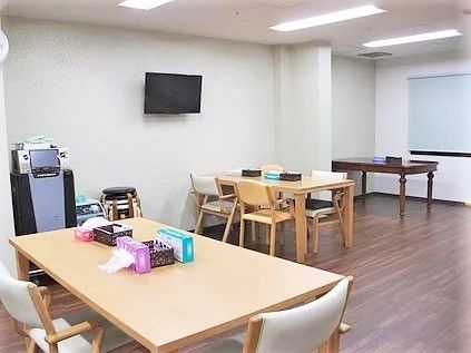 フローリングのリビングダイニング。木製のテーブルが複数置かれ、壁掛けテレビもある。