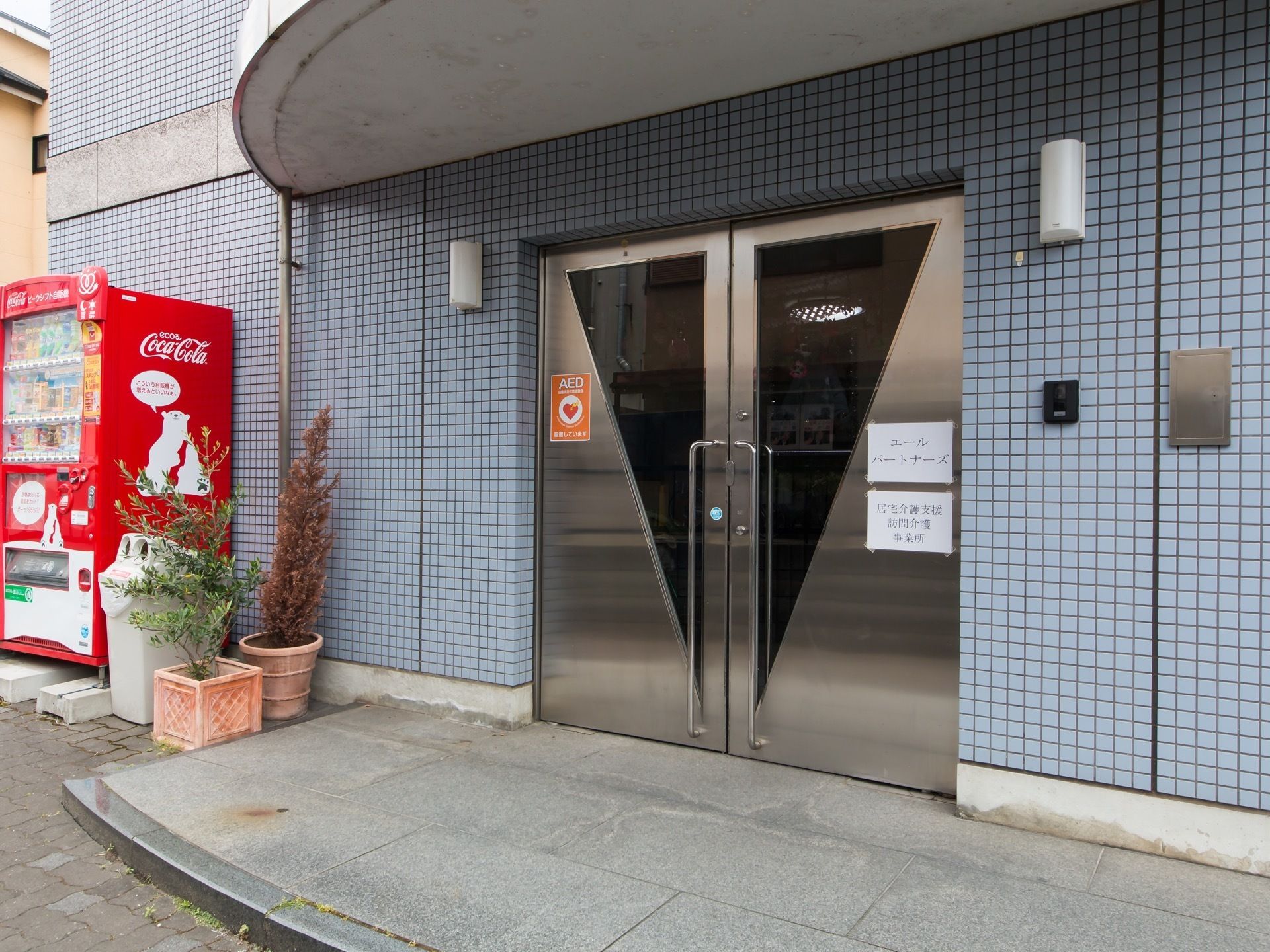 青い細かいタイルの壁の玄関先。金属製の扉があり、半円形のルーフが付いている。傍らには観葉植物と自動販売機が置かれている。