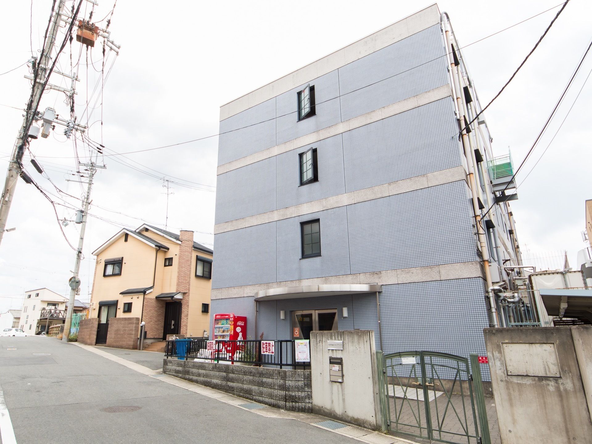 青いタイルで覆われている４階建ての大きな建物がある。道路沿いに面しており、コンクリートブロックとフェンスで仕切られている。