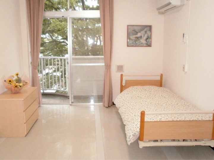 ベランダ付きの居室には寝具付きのベッドがあり、花が飾られたタンスが置かれ、明るい雰囲気。