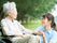 緑に囲まれた屋外で過ごす眼鏡をかけた高齢女性とブルーのポロシャツを着た若い女性スタッフ。女性は車いすに座っている。