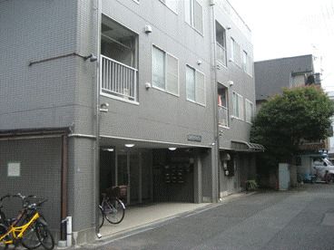 グレー系の3階建ての建物の玄関の前には自転車が止められている。窓が多く付いたホーム。