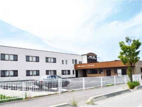 白い外壁の２階建ての建物は日当たり良好で一部ブラウン系の建物が併設されている。駐車場もある。