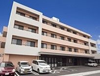 駐車場付きの4階建ての建物は横に長く、ベランダも付いている。駐車場には数台の車が止められ、車いすマークも描かれている。