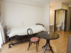 寝具付きのベッドが壁際に置かれ、円形のテーブルセットが置かれている。奥にはトイレがあり、トイレのドアが開いている。