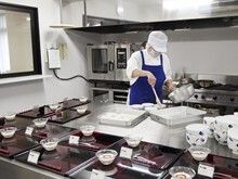 青いエプロンを付けたスタッフは白い帽子とマスクを付け、料理を器に注いでいる。周りにはたくさんのトレーが置かれている。