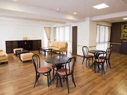 大きな窓がたくさん付いた共有スペース。手前には円形のテーブルセットが二組あり、奥にはソファの応接セットが置かれている。