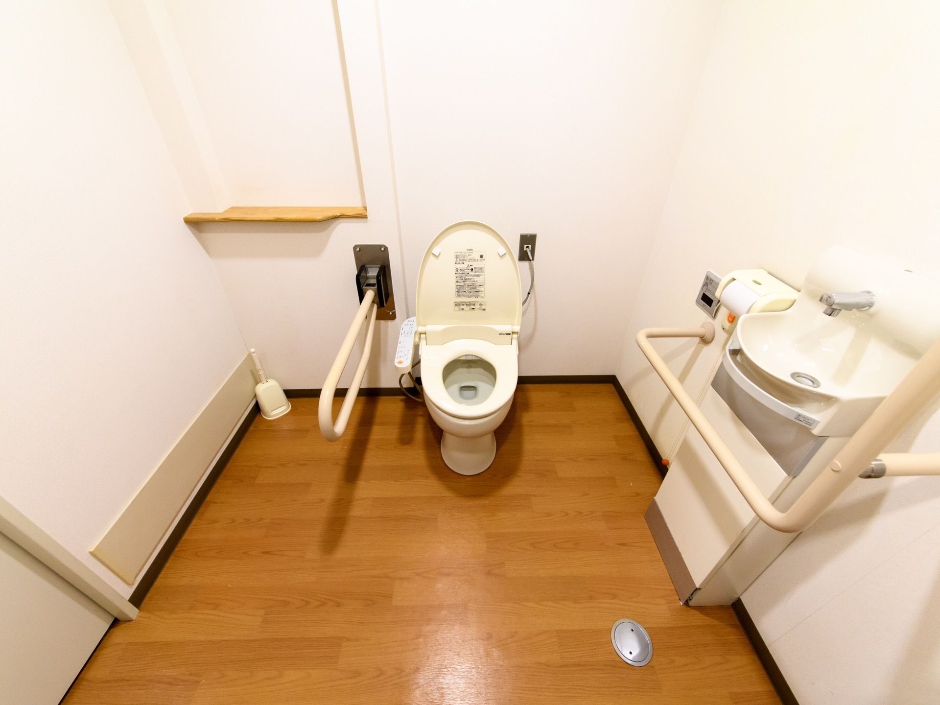 木目調の床に白い壁面の内装である。手洗いスペースと便器付近に手すりが設置されている。緊急呼出用のボタンがある。