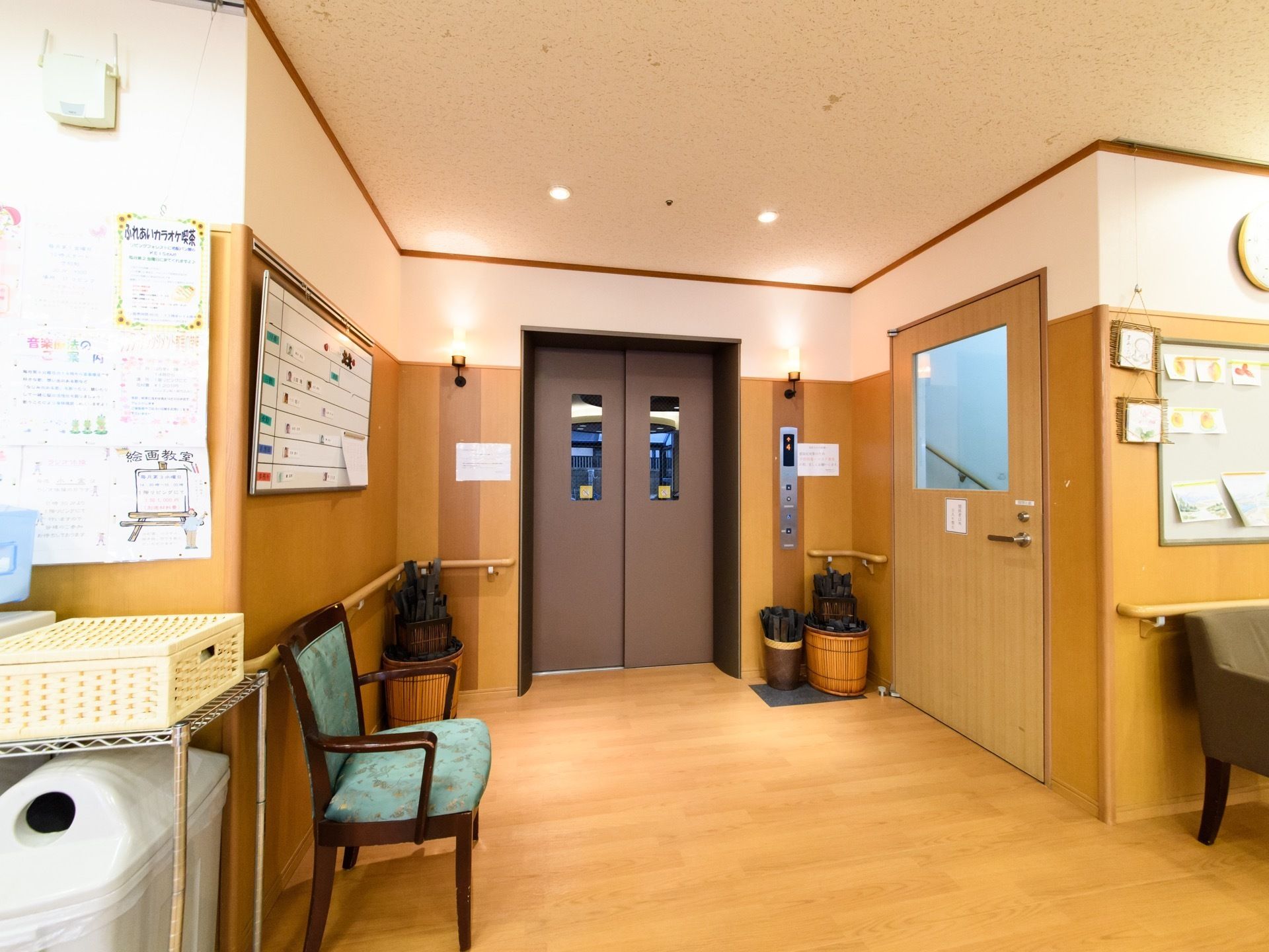 床は木目調である。エレベーターは車椅子対応の機種である。イスが設置してある。壁にはイベントのお知らせが貼ってある。