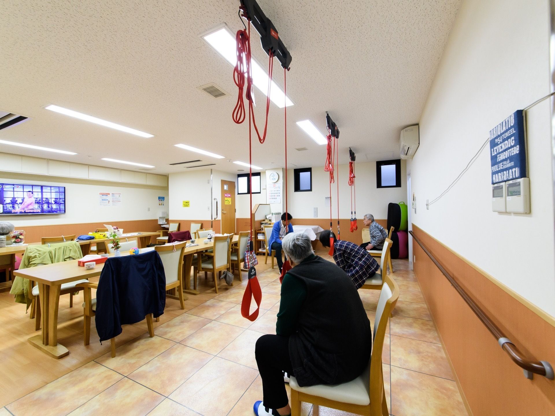 大きな液晶テレビが1台置かれている。長テーブルの周りには背もたれ付きの椅子があり、近くでは機能訓練が行われている。