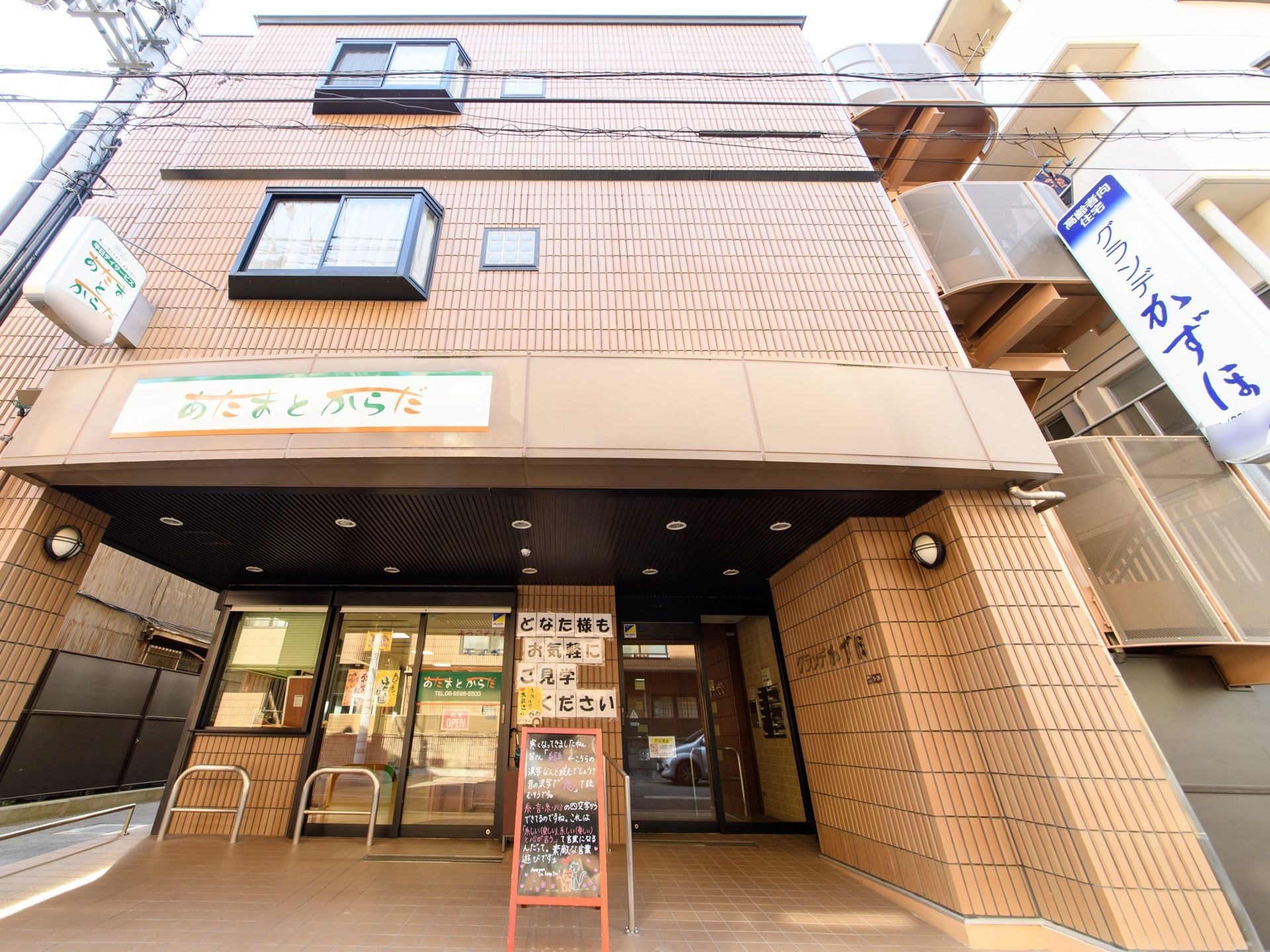 3階建ての茶色いタイルの建物がある。1階はガラス張りになったデイサービスがある。隣には高齢者向住宅がある。