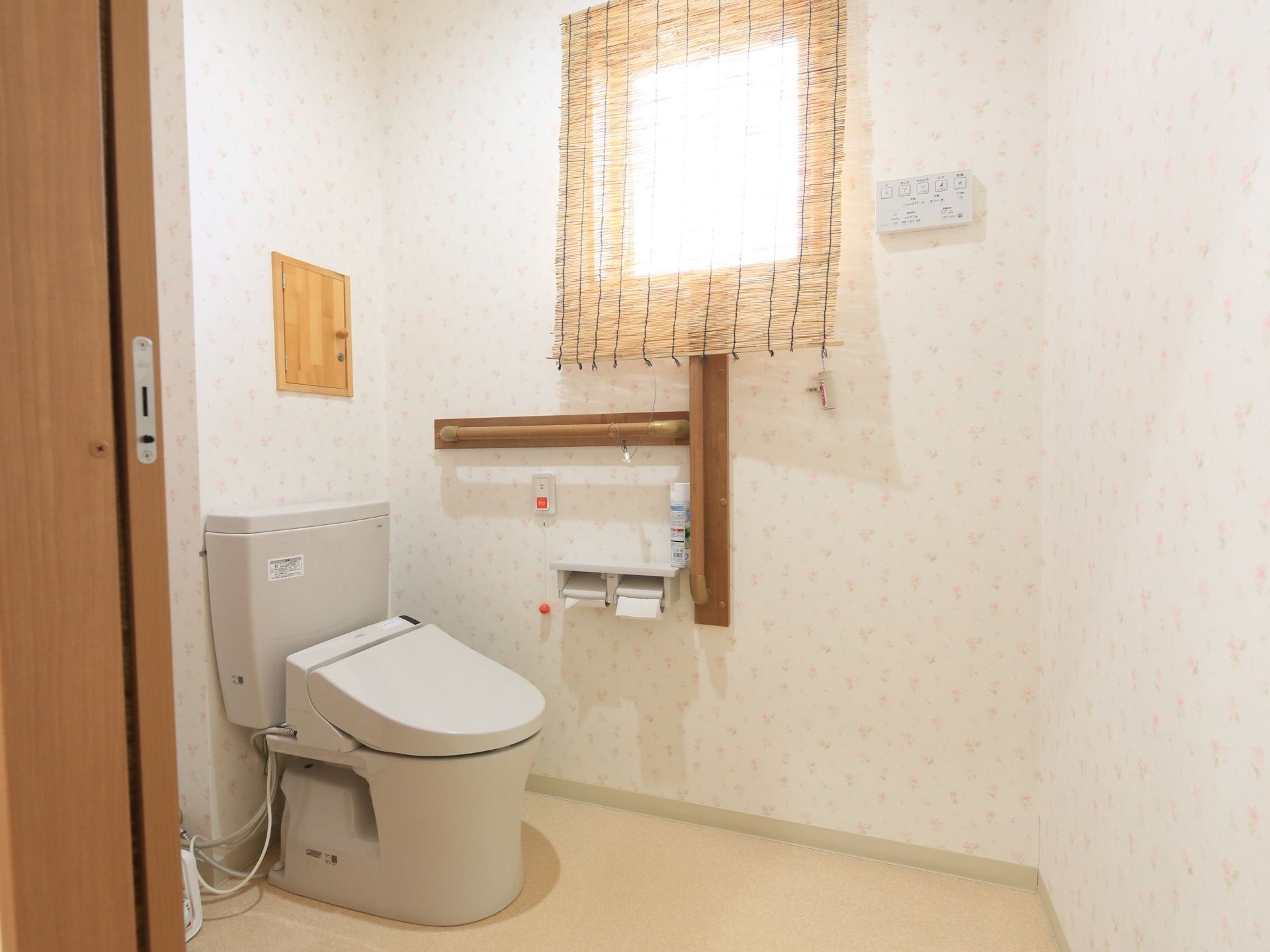 花柄の壁紙が貼られた個室トイレである。温水洗浄便座が設置されている。窓にはすだれがかけられ、日よけがされている。