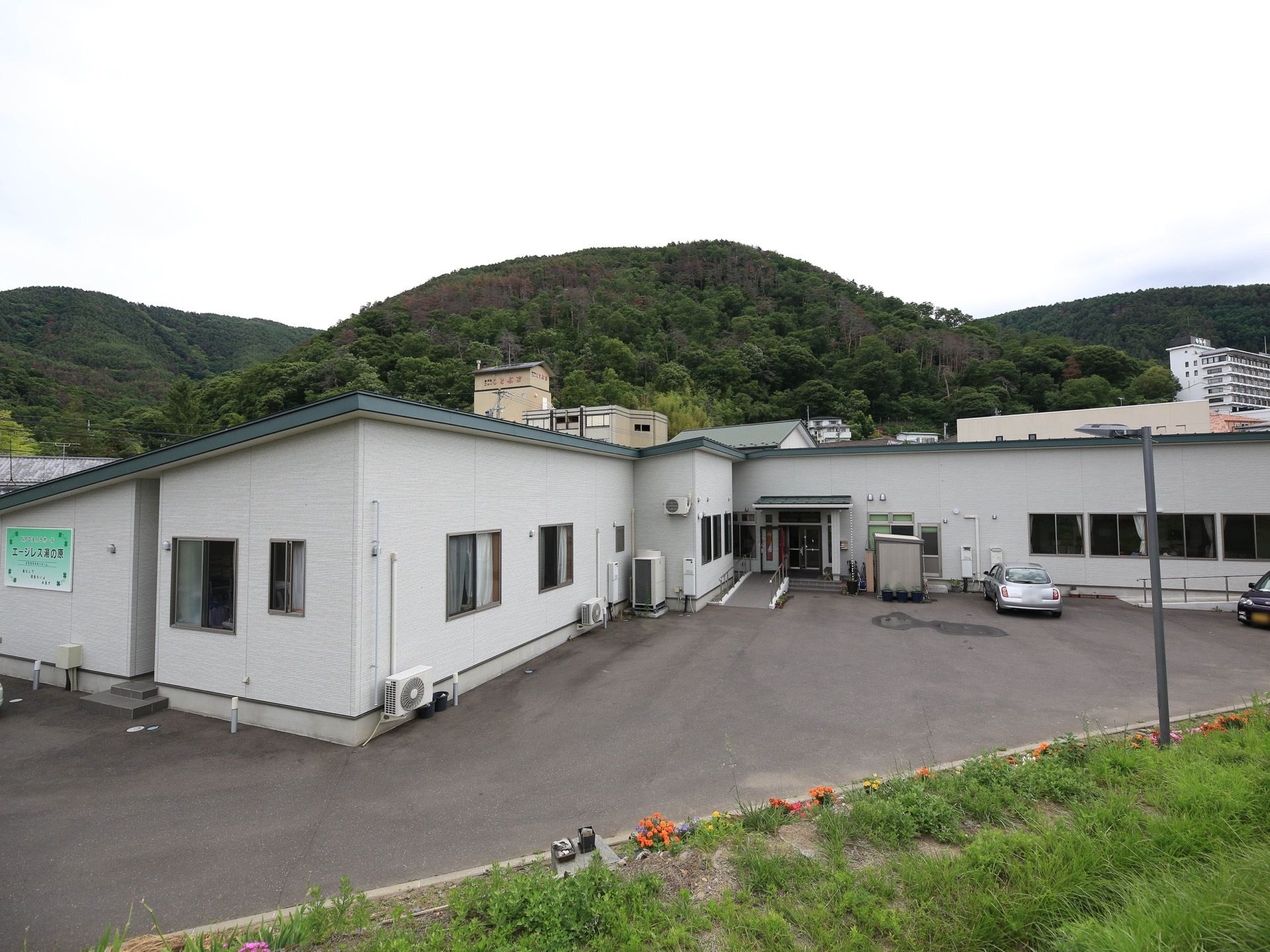 白い外壁の施設で、建物の前は駐車スペースになっている。施設の裏側は小高い山がそびえており、自然に溢れている。