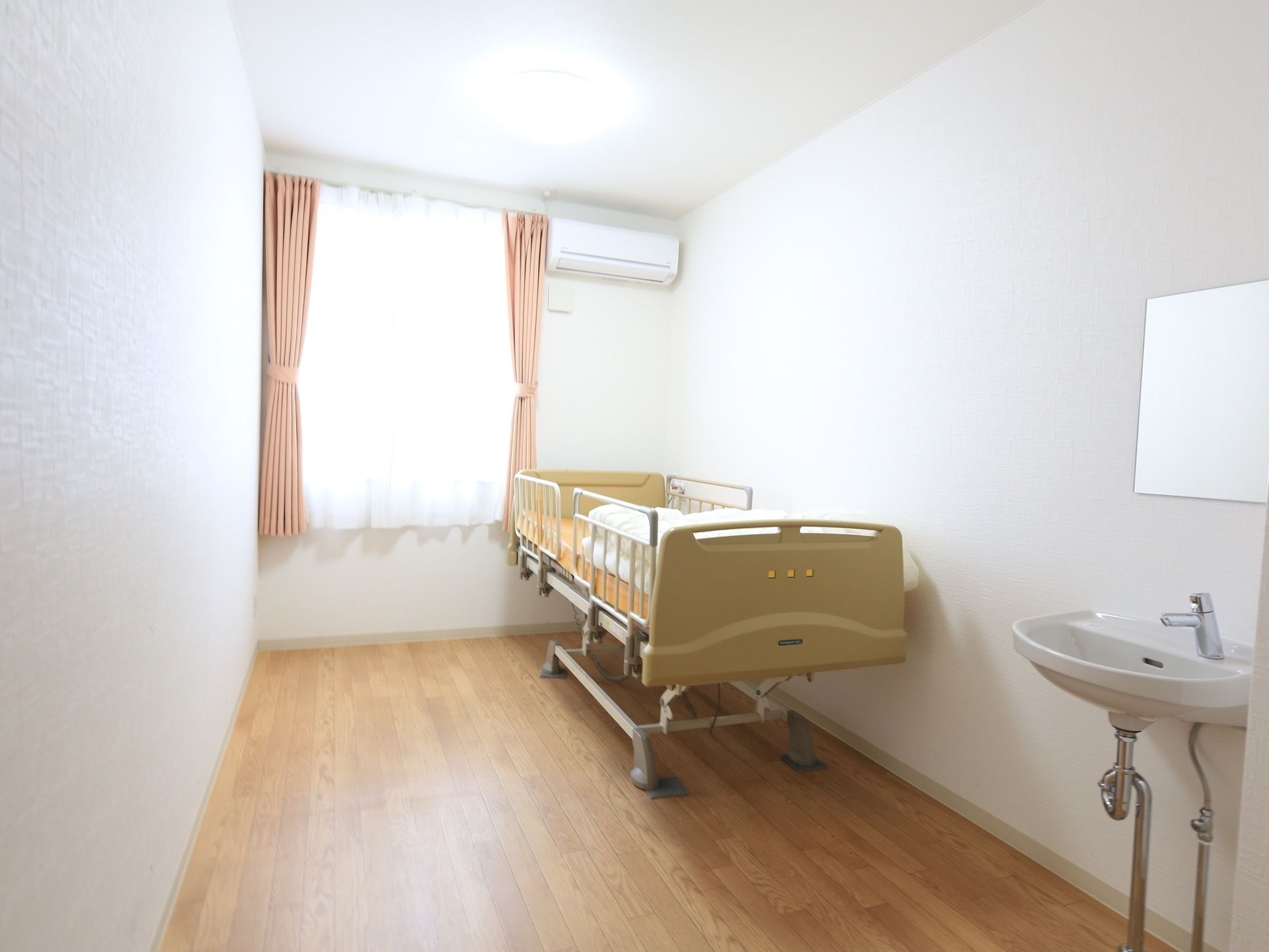 板張りの床に白い壁の居室で、大きな窓にはピンクのカーテンが掛けられている。壁際に介護用ベッドが置かれている。