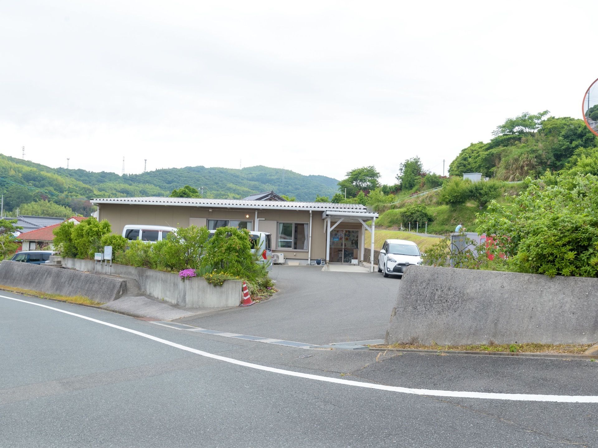 建物周りに、木や植物が複数植えられた場所である。数台の車が敷地内に停まっており、道路にはカーブミラーが設置されている。