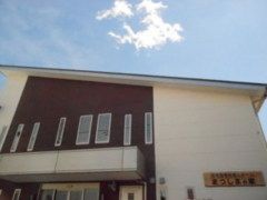 建物外観のアップ。青い空の下に白と茶色の建物がある。玄関の上には屋根が付いている。