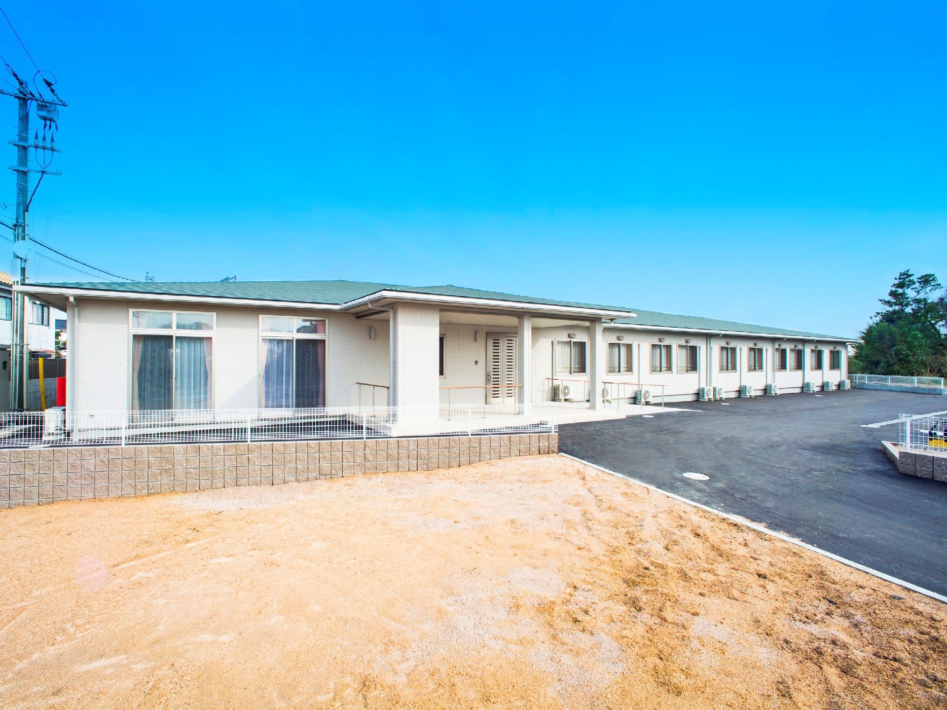 平屋建てのベージュの建物。白いフェンスで囲まれ、広い駐車場もある。