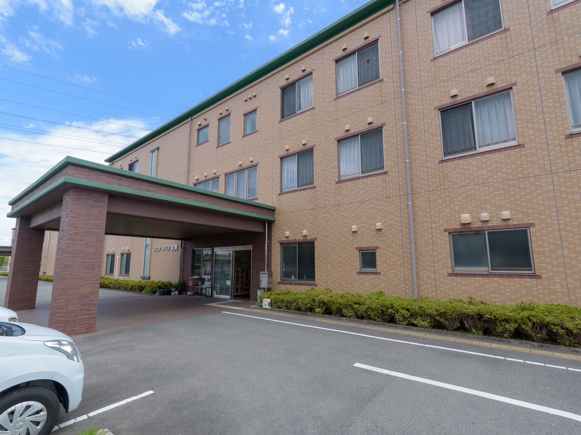 青空が広がり、茶系の３階建ての建物がある。窓が規則的に並んで設置されており、建物前には駐車スペースがある。