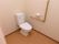 広いトイレに白い洋式便器が設置されている。壁にはＬ型の手すりが設置され、ペーパーホルダーの横にナースコールがある。