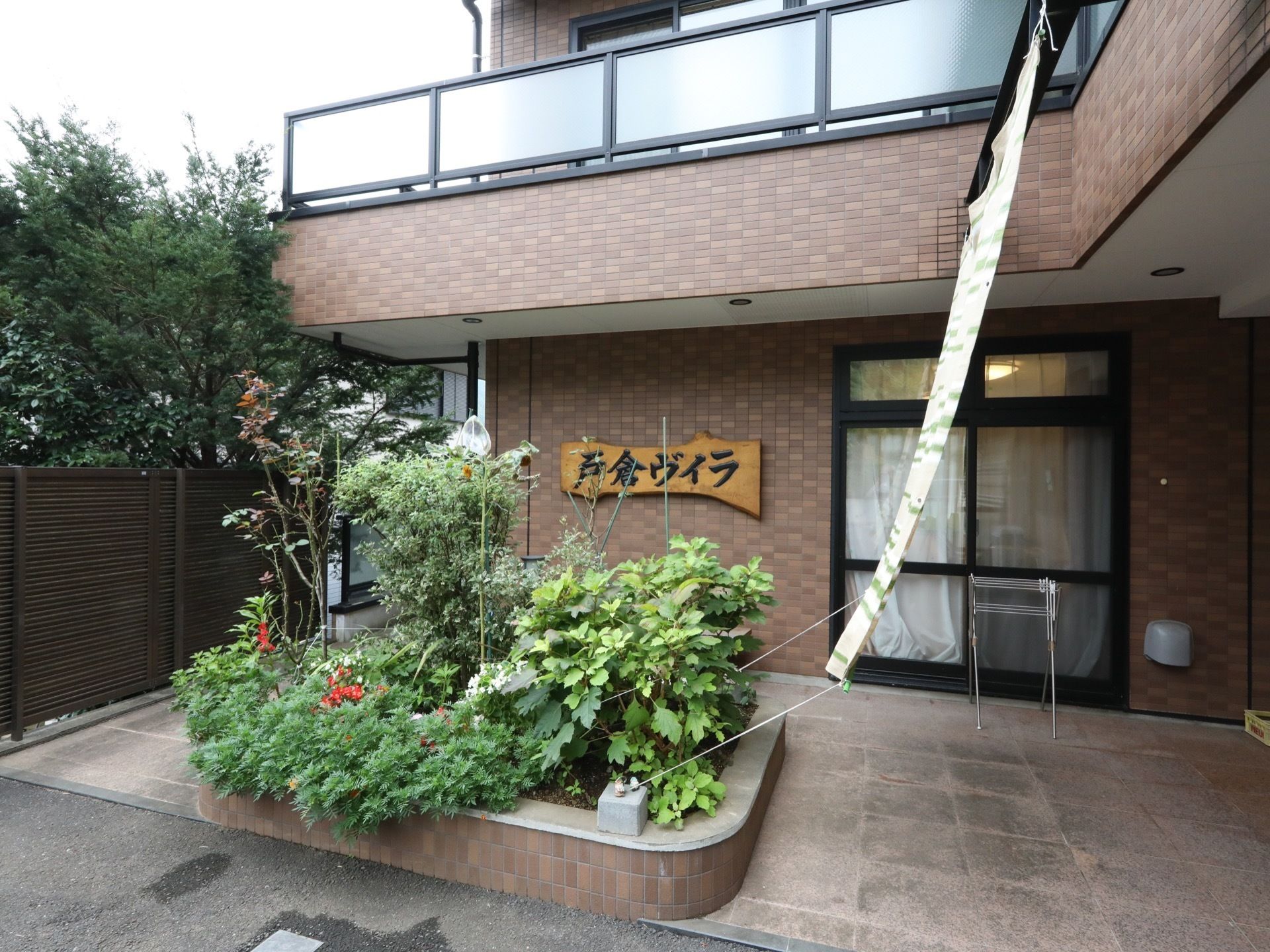 茶色いタイル張りの外壁に、樹木の形状を感じられる材木を用いた施設名表示板がある。植木や草花が密集している花壇がある。