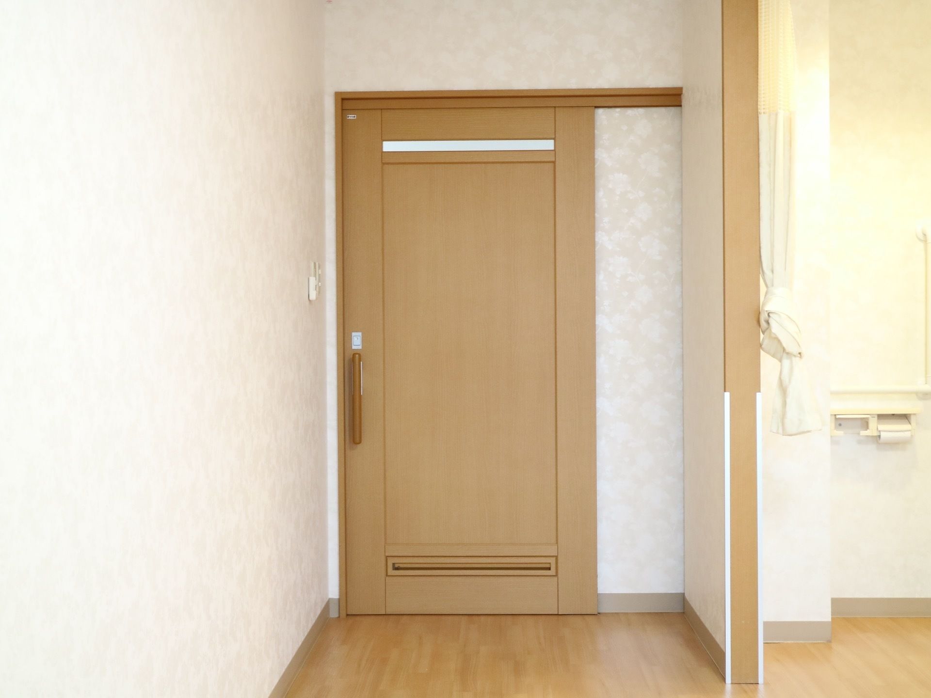 白地に光沢のある花柄が描かれた壁紙が施されている室内。正面に木目調の扉が設置されており、右側のトイレの壁にペーパーホルダーがある。