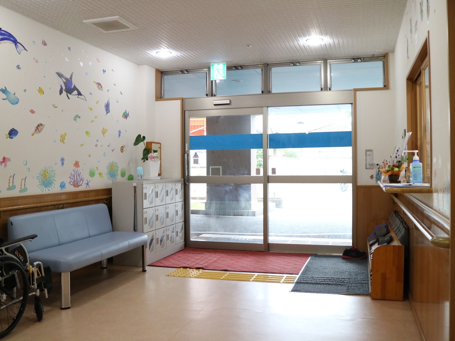 大きなガラス製の引き戸が設置されている玄関である。左側に下駄箱とベンチ、右側にスリッパラックがある。事務所の窓口が設けられている。
