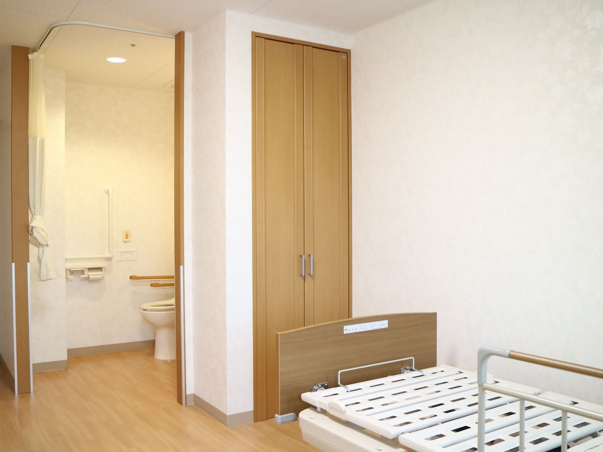 天井にＬ字型のカーテンレールが設置されており、その奧に洋式便器が据えられている。ベッドのそばにクローゼットの扉がある。