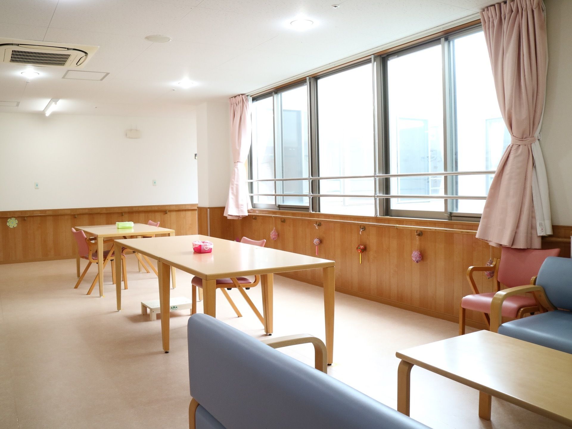 窓が並ぶ広間に、木製のテーブルや椅子が設置されている。手前には水色のソファが置かれているのが見える。