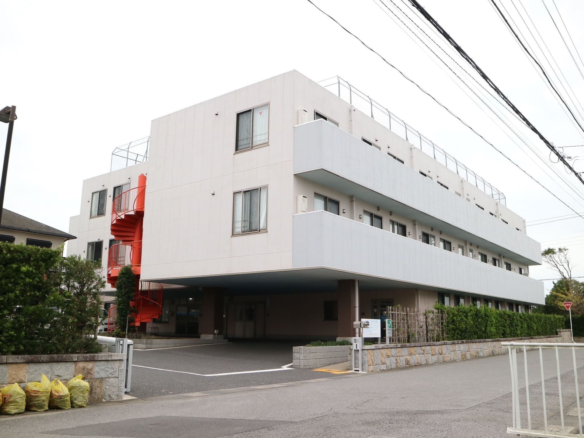 公道沿いに建つ白い３階建ての施設。生け垣が施されており、広い間口の奧には舗装されたスペースが広がっている。