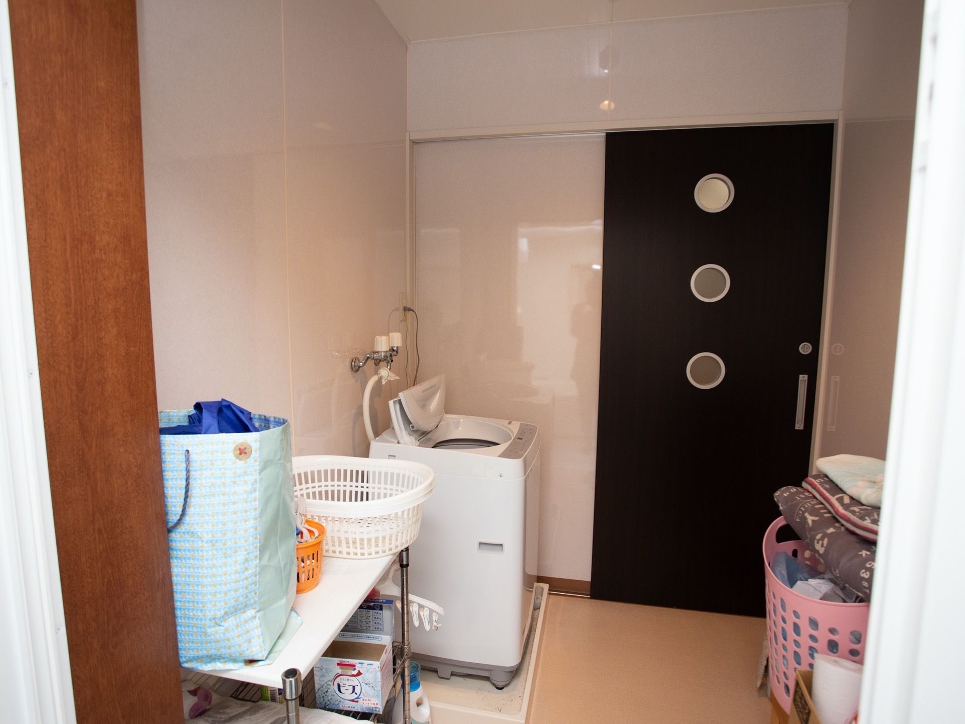部屋の片隅に洗濯機が１台設置され、横に簡易棚がある。棚の上には脱衣かごや紙袋が置いてあり、向かい側の脱衣かごには衣類が置いてある。