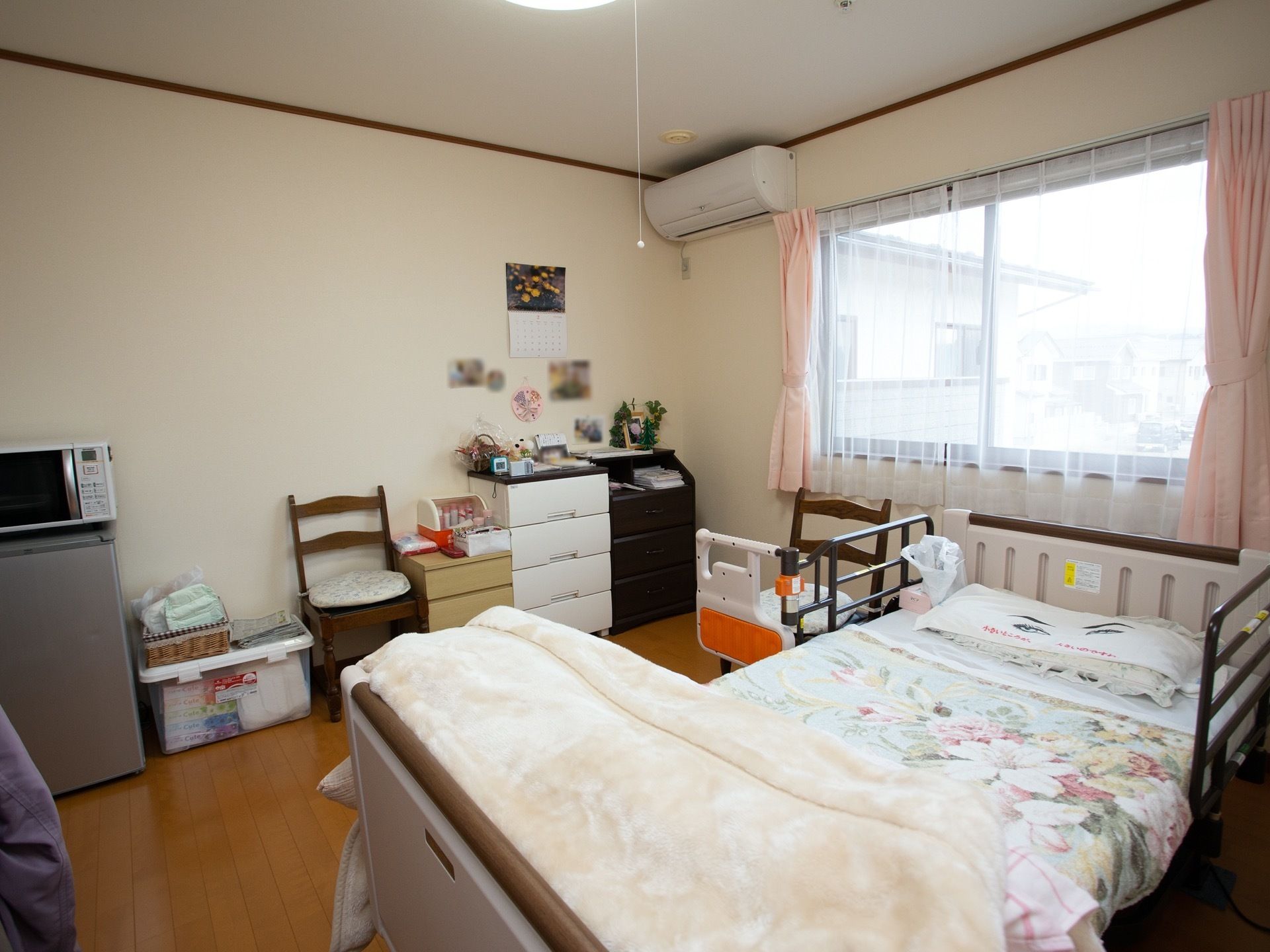 居室は白を基調とした壁紙、木目調のフローリングというデザインである。ベッドのほか、複数の収納棚や椅子、電子レンジなどが置かれている。
