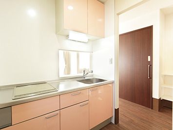フローリングの部屋にはサーモンピンクの収納扉が付いたIHクッキングヒーター付きのキッチンが付いている。