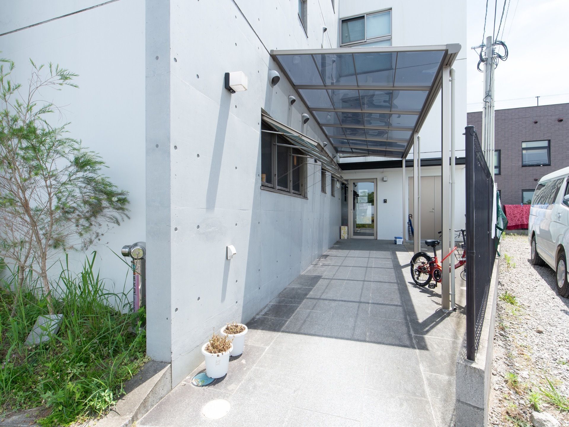 正面に、ホームの玄関に続く通路があり、通路の上方には透明感のある素材の屋根が備え付けられている。また通路の横には、駐車場も設置されている。