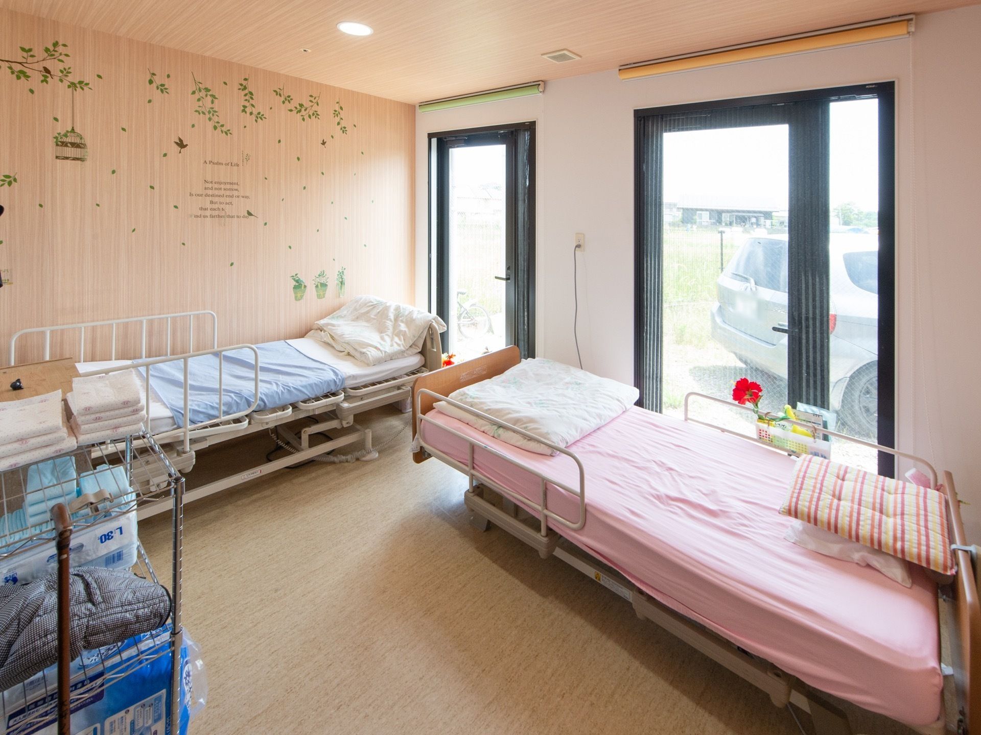 大きな窓が２つあり、介護用ベッドが２台設置されている居室。壁には、木や鳥などのイラストや、英文などが書かれている。