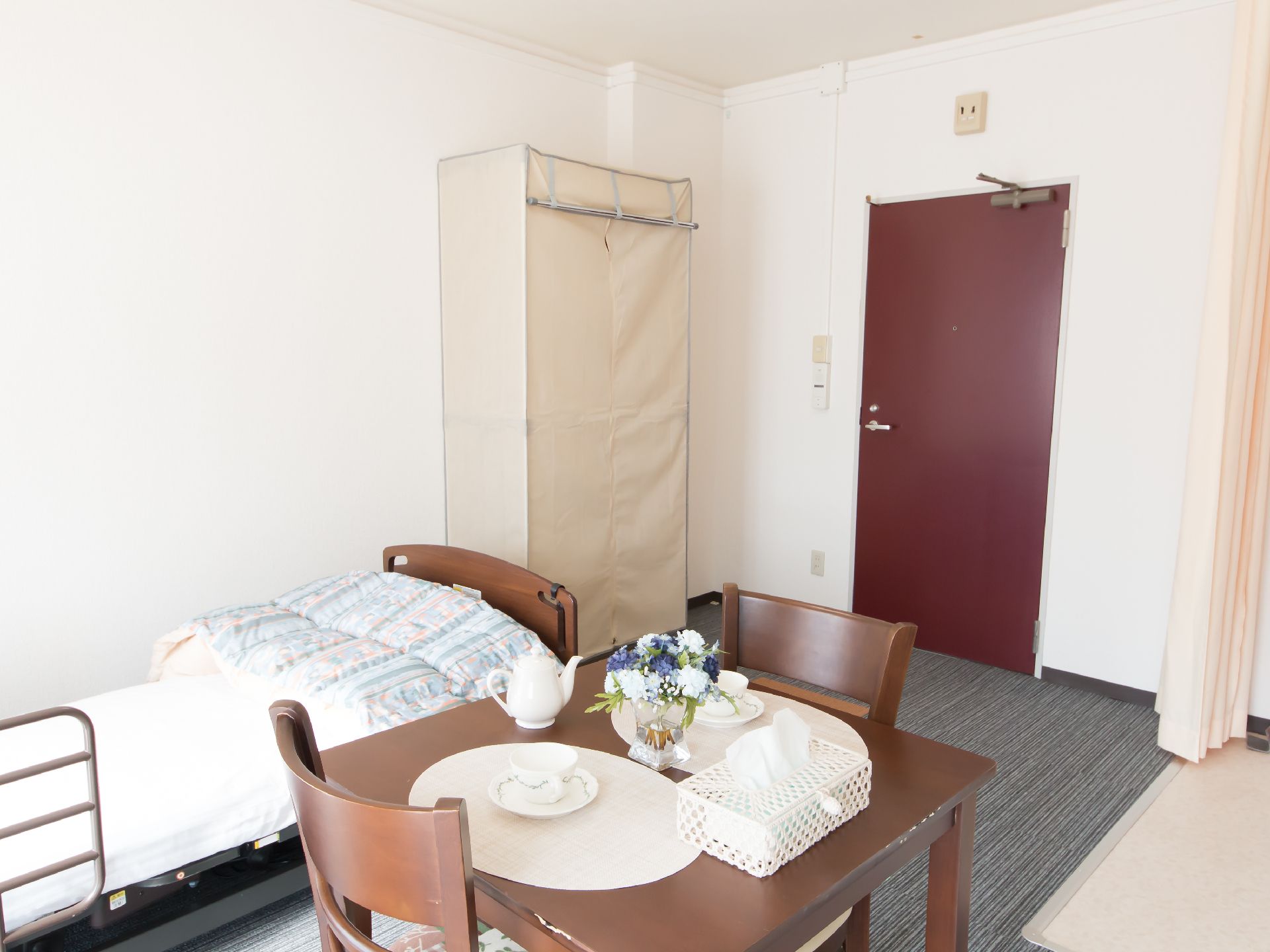 居室の入口は、濃い赤色の開き戸になっている。転落防止柵つきのベッドやダイニングテーブルが設置されている。