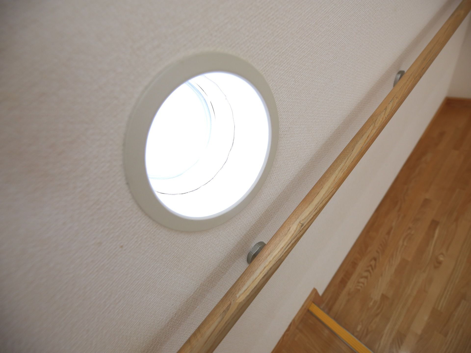白い壁に木製の手すりと円形の小窓がある。外の明るさが分かり、壁には手すりの影ができている。