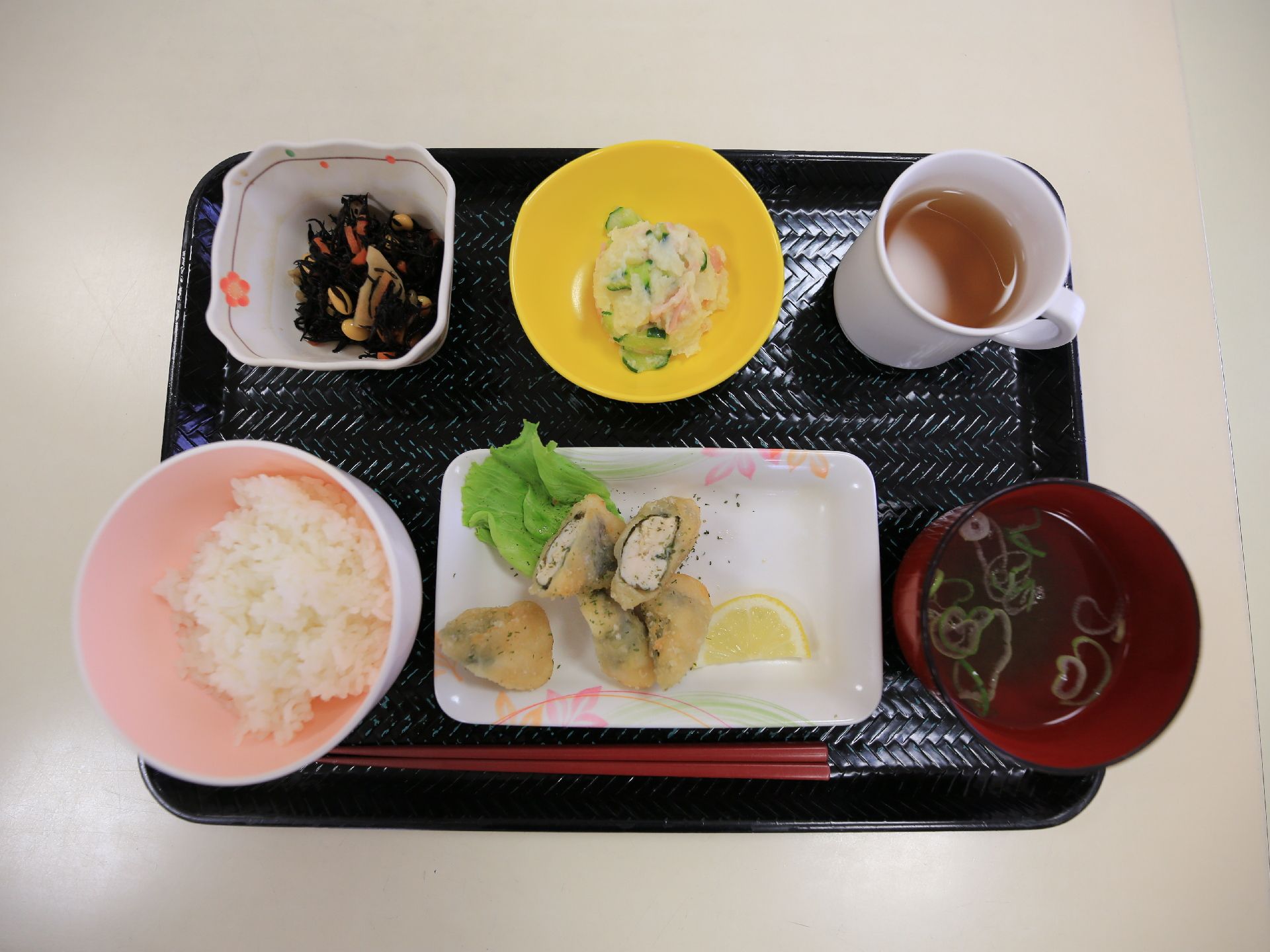 ある日の一汁三菜の献立である。それぞれ器に盛り付けられて、黒いプレートの上に赤いお箸や飲み物と共に並べられている。