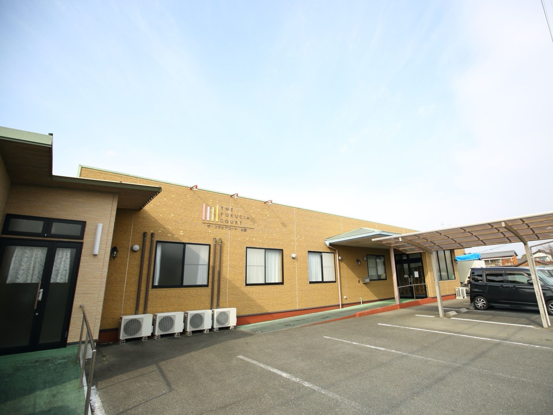 明るいブラウン調の外観である。外壁に施設名やマークが描かれている。エントランスから駐車場にかけて、屋根がある。