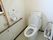 アイボリーの洋式便座が備え付けられた、トイレが完備されている。壁紙はホワイト調。木製の手すりが設置されている。
