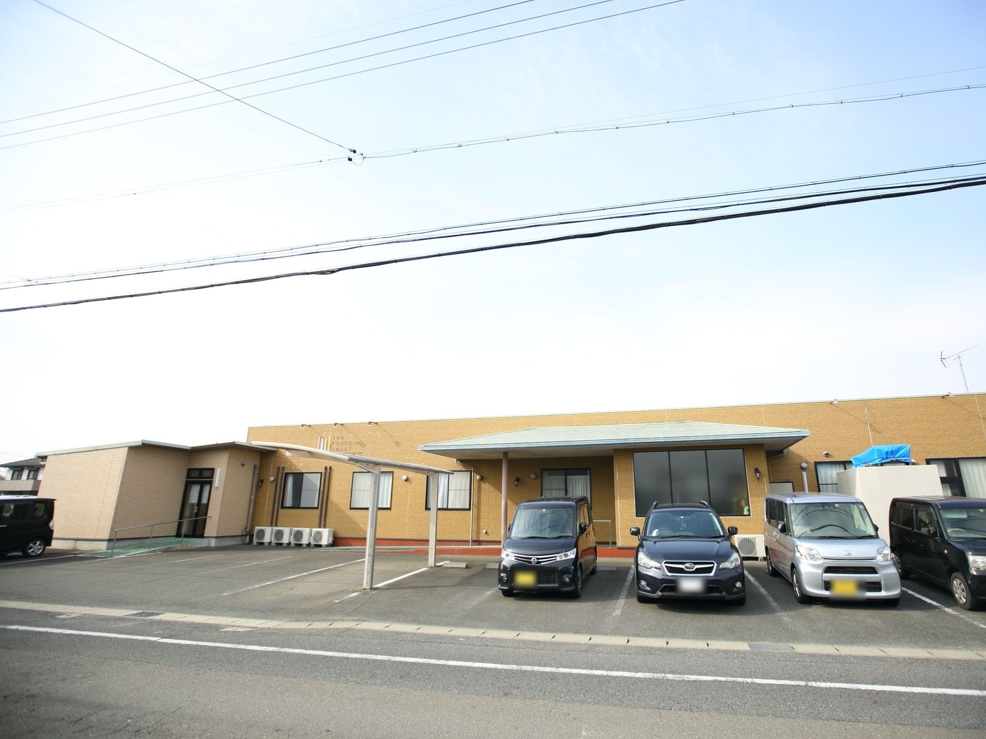 道路に面したところに建つ、平屋建ての施設である。外壁は明るい色合い。駐車場には複数台の車が停まっている。