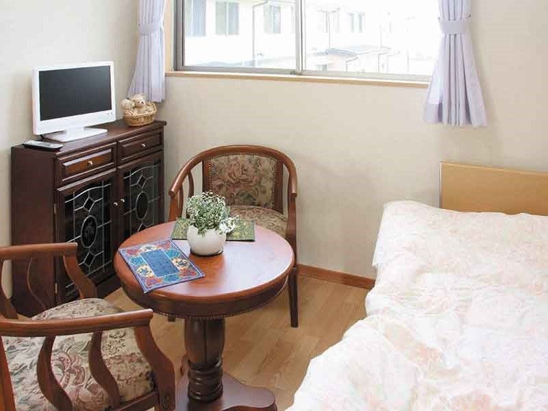 フローリングの居室にはエアコンやカーテン付きの窓があり、ベッドやテーブルセット、テレビなども置かれている。