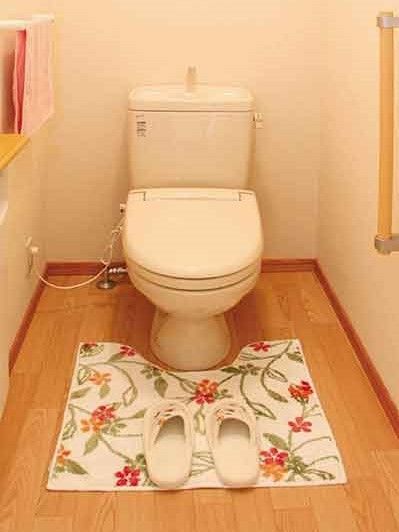 温水洗浄便座のシンプルなデザインのトイレ。アイ字型の手すりもあり、花柄のマットとスリッパもある。